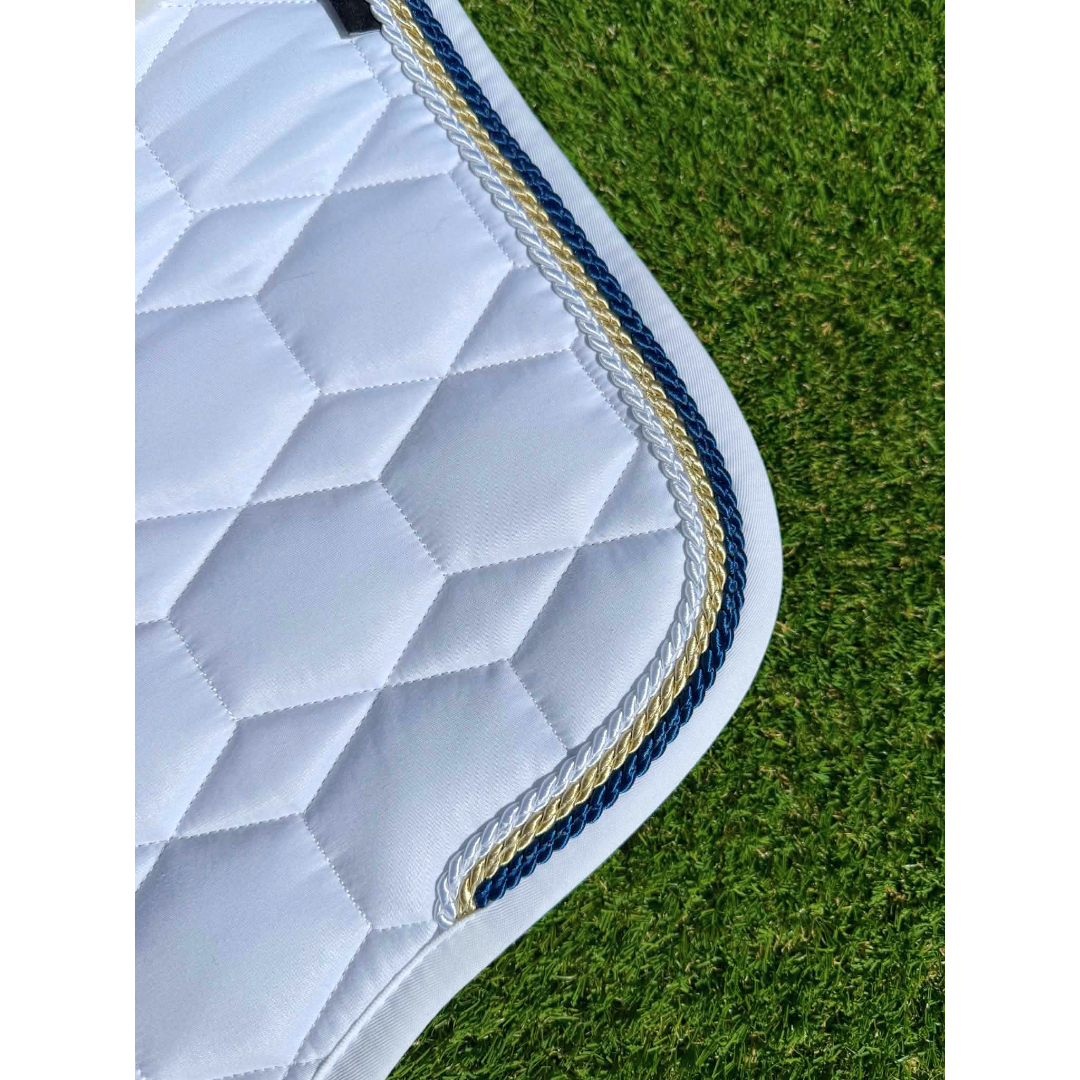 A close-up of a white quilt sheen horse saddle pad with a decorative piping of steward, champagne, and white, laid on faux green grass background.